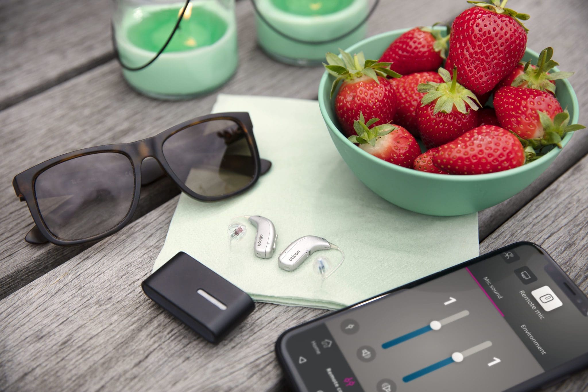 A pair of sunglasses, hearing aids, a smartphone with an app open, and a bowl of strawberries are placed on a wooden surface with a green napkin.
