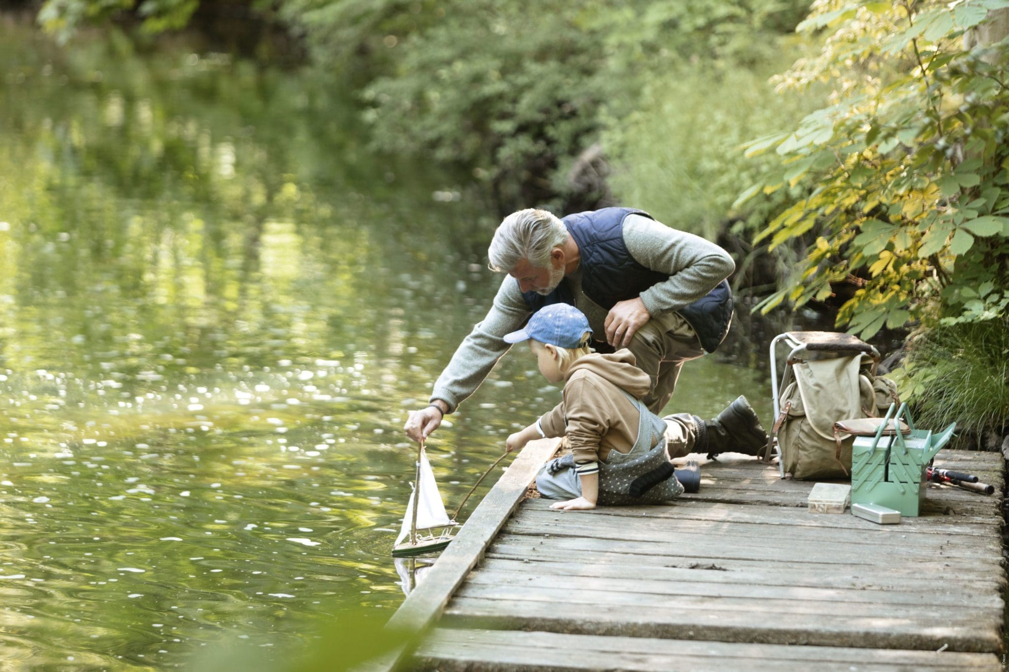 An adult and a child are crouching on a wooden dock by a pond, launching a small toy sailboat into the water. Fishing and camping gear are nearby.
