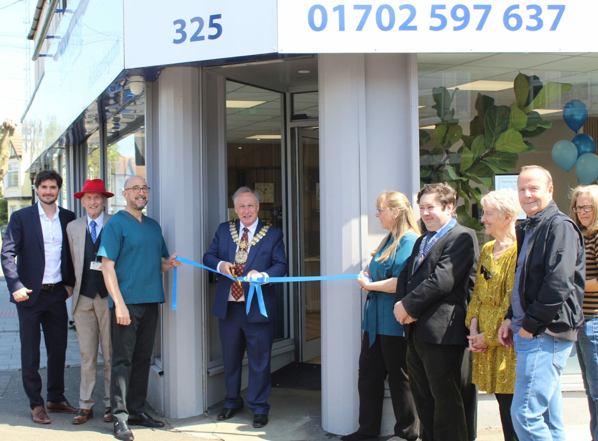 A group of people, including a man in regalia with a ceremonial chain, stand outside a building during a ribbon-cutting ceremony.