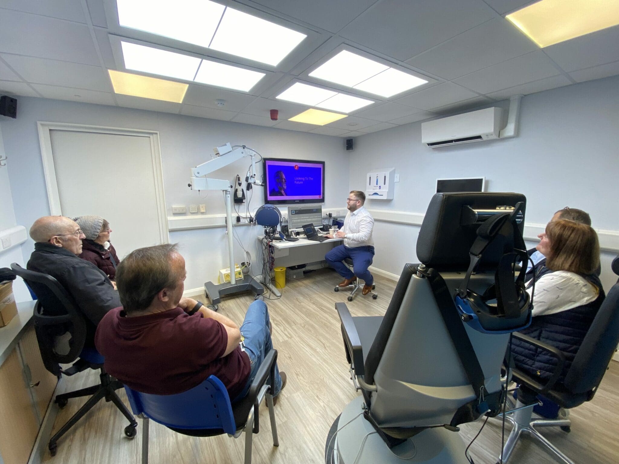 A group of people are sitting in a medical office listening to a presentation from a person at a desk with a computer screen. Medical equipment is visible in the room.