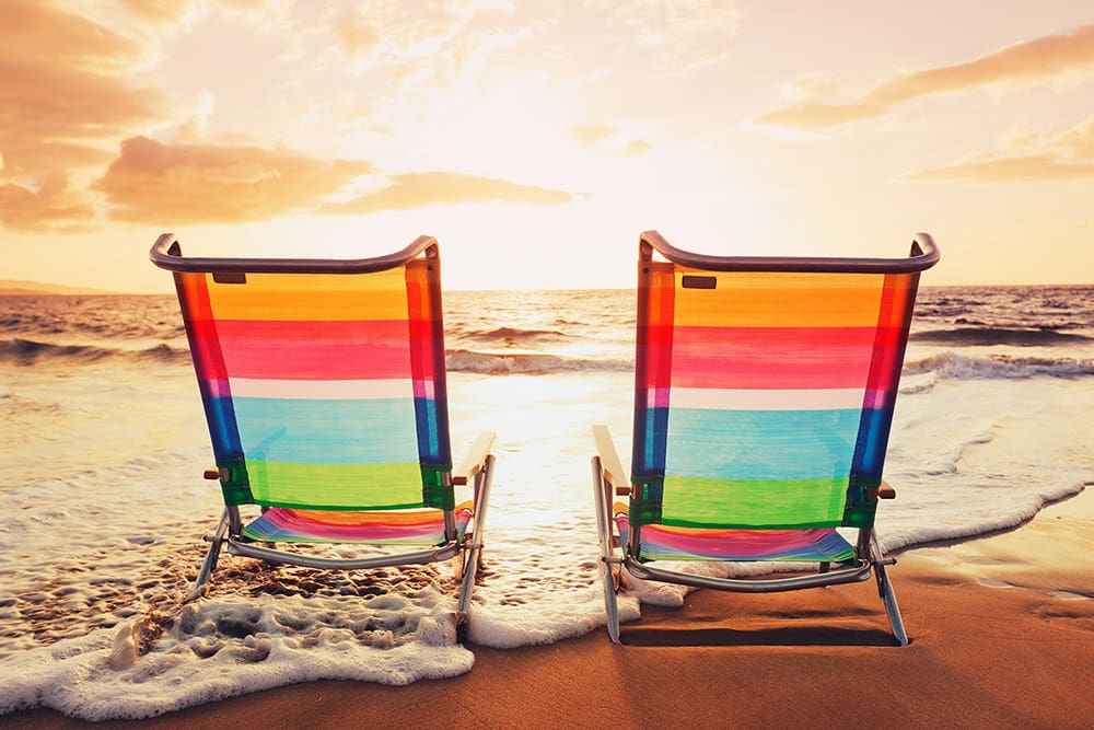 Two colorful beach chairs sit on the sand facing the ocean, with gentle waves washing up and the sun setting in the background.