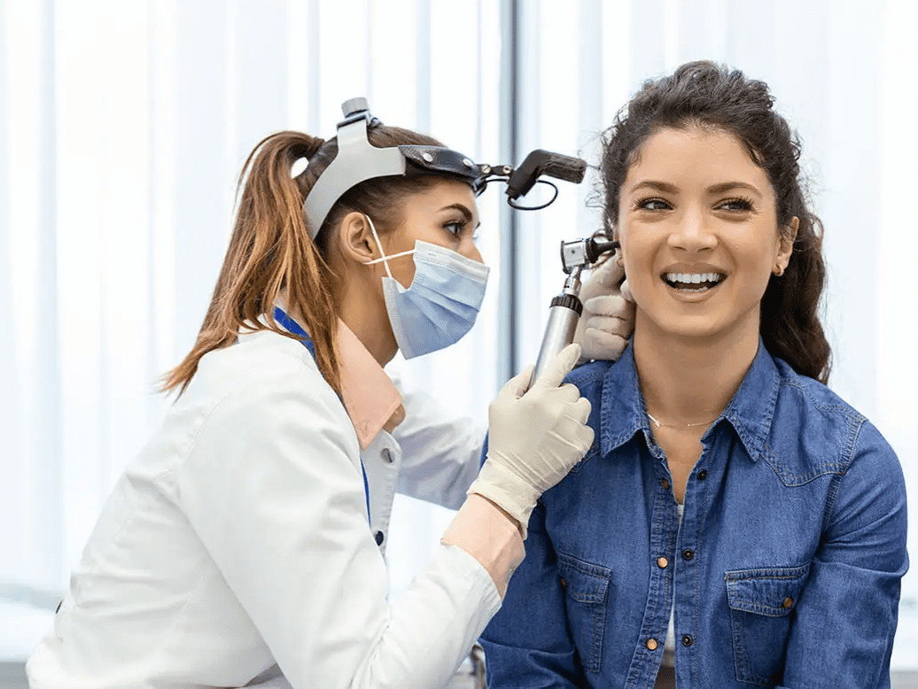 A healthcare professional wearing gloves and a mask examines a smiling woman's ear with an otoscope in a medical office.
