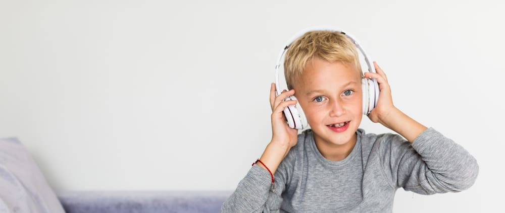 Young boy with blonde hair wearing a gray shirt and white headphones, sitting on a couch and smiling at the camera against a plain white background.