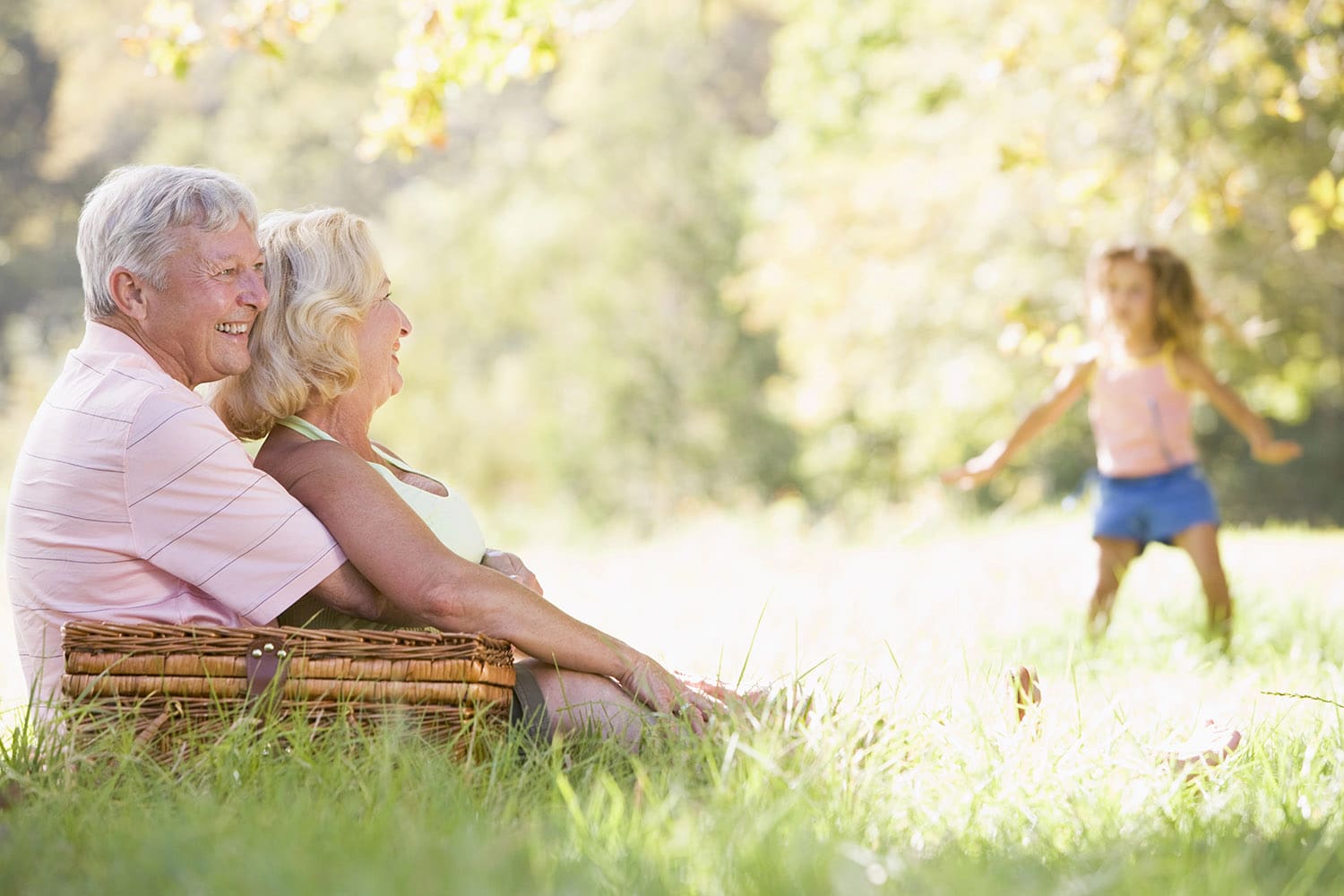 Two older adults sit on the grass with a picnic basket, smiling and watching a young child run toward them in a sunny outdoor setting.