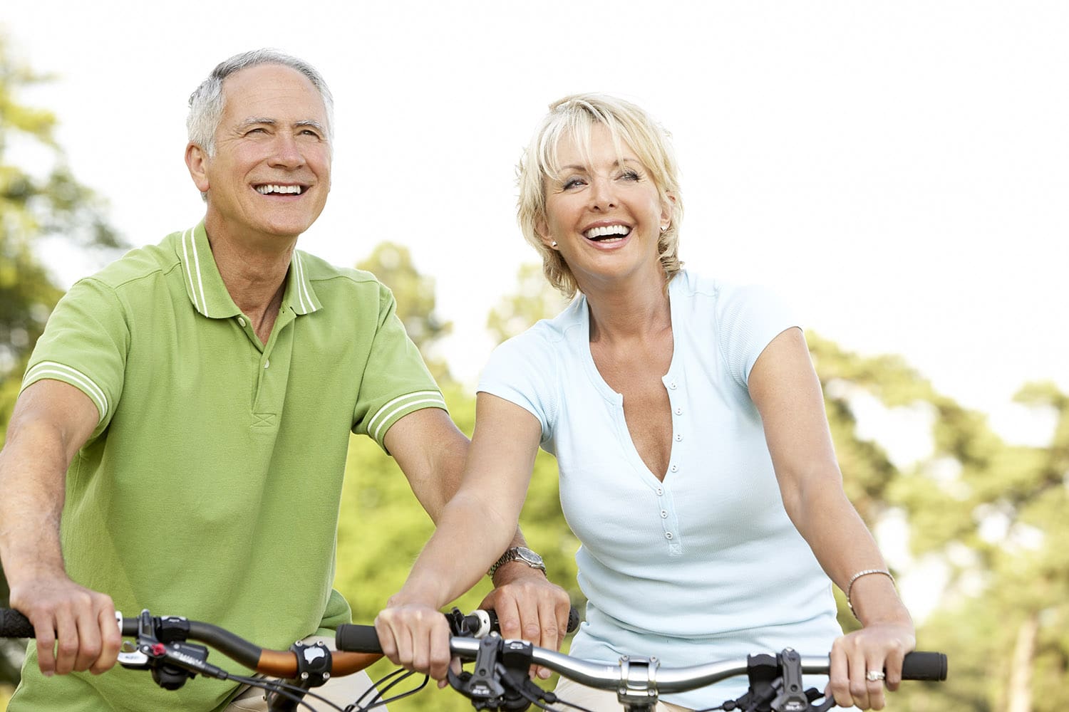 Two smiling older adults, a man and a woman, ride bicycles outdoors on a sunny day, surrounded by green trees.