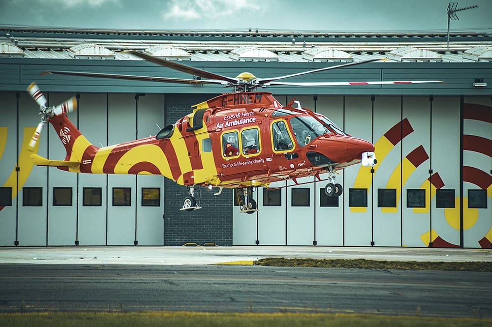 A red and yellow air ambulance helicopter hovers above the ground in front of a hangar with matching red and yellow designs.